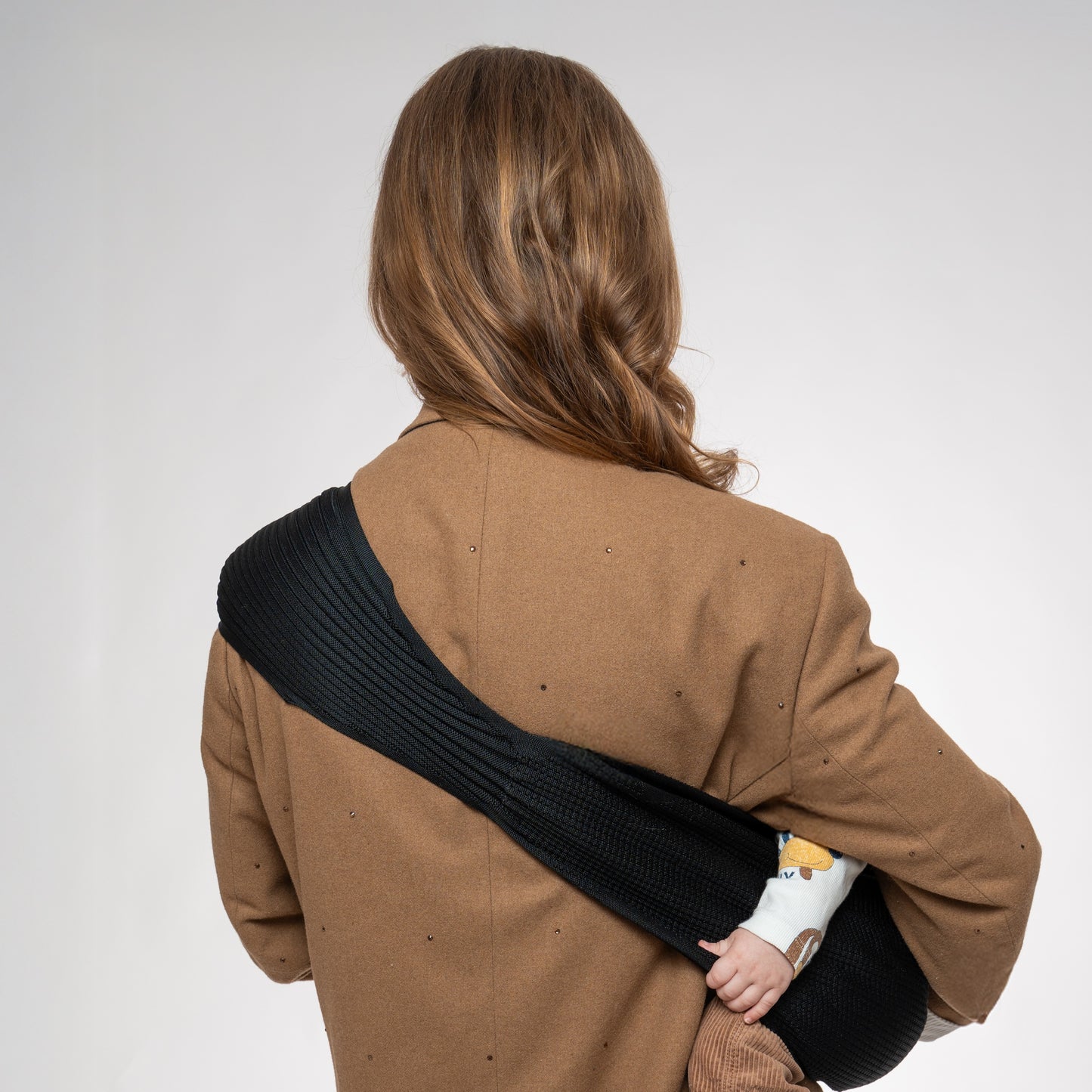 Rear view of a woman wearing a blazer, highlighting the wide ergonomic shoulder support of the black Coshen 3D knit carrier. The accordion-style knit distributes weight evenly to prevent neck and shoulder strain during long-term use.