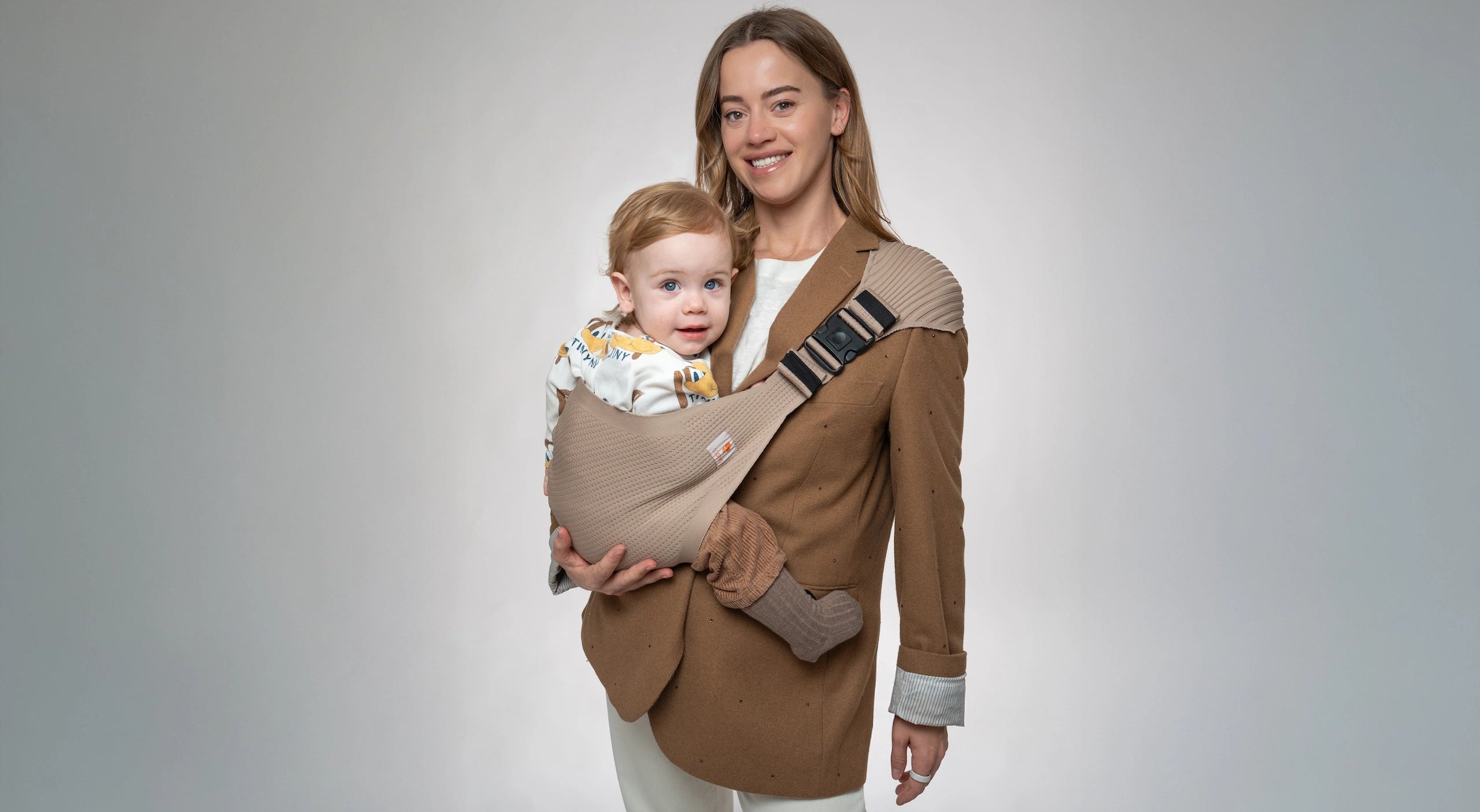 Brede collectiebanner van een lachende moeder in een bruine blazer die de beige Coshen 3D-knit peuter heupdrager stijlvol gebruikt; de ultieme ergonomische en premium draagoplossing voor moderne ouders.