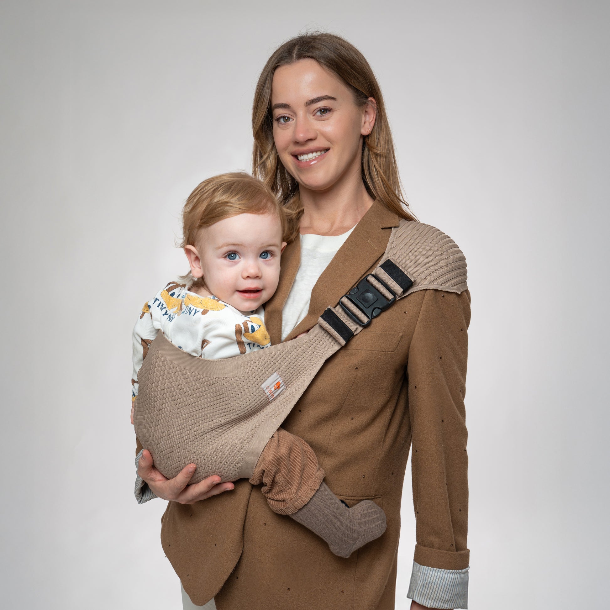 Front view of a woman wearing a brown blazer using the Coshen seamless 3D knit toddler hip carrier in beige. The image showcases the ergonomic M-position for the baby and the stylish, minimalist design for modern parents.