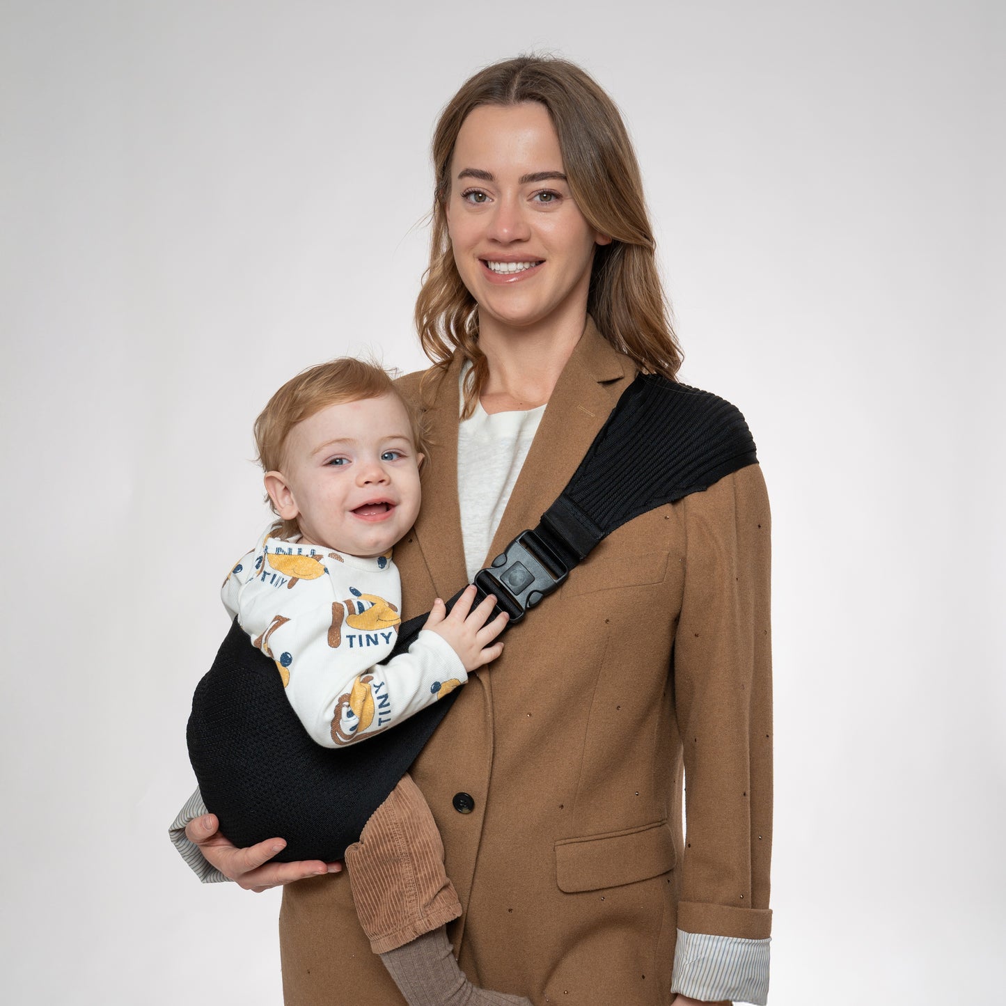 Front view of a woman wearing a brown blazer carrying a toddler in a black Coshen seamless 3D knit hip carrier. Showcases the ergonomic M-position for hip health and a stylish, minimalist look for modern parents.