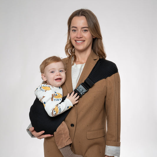 Front view of a woman wearing a brown blazer carrying a toddler in a black Coshen seamless 3D knit hip carrier. Showcases the ergonomic M-position for hip health and a stylish, minimalist look for modern parents.