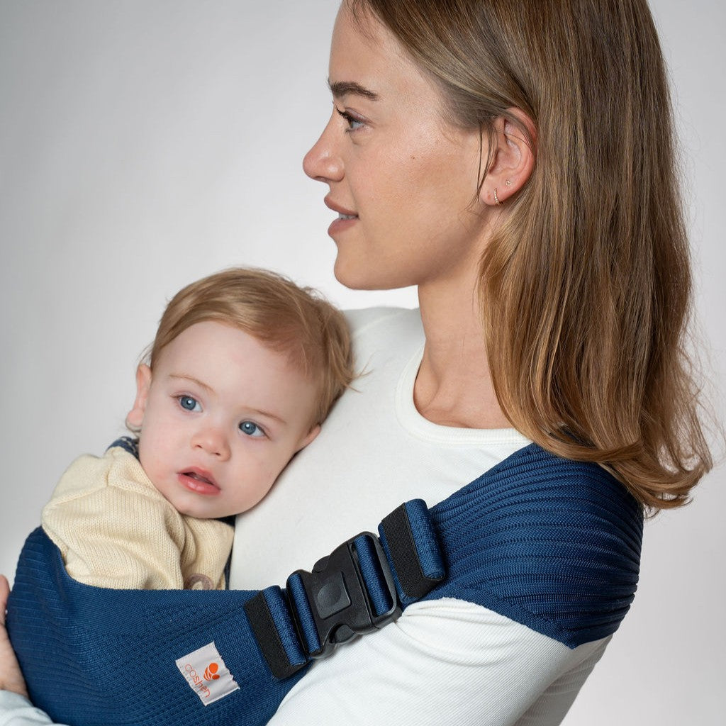 Side profile view of Coshen navy blue seamless knit toddler hip carrier with a baby, showing the buckle close-up and white long-sleeve shirt.