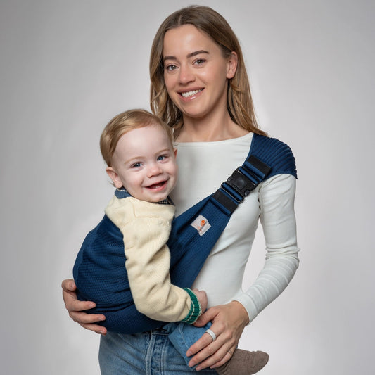 Smiling woman wearing a navy blue seamless knit Coshen toddler hip carrier with a happy baby facing forward.