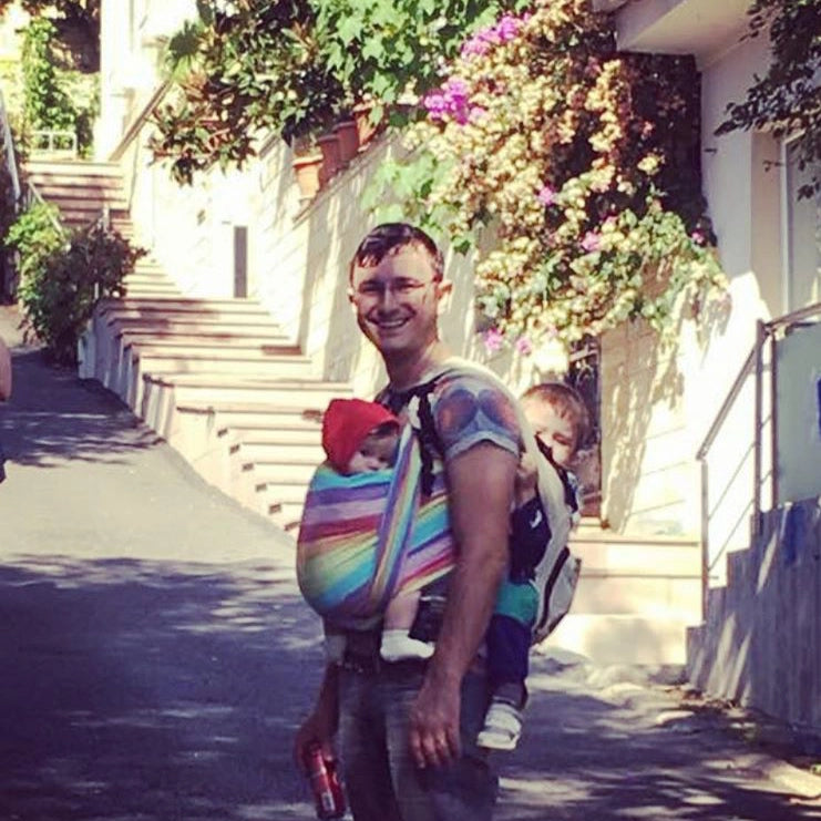 A smiling father and baby carrier manufacturer carrying two small children in a tandem carry outdoors.