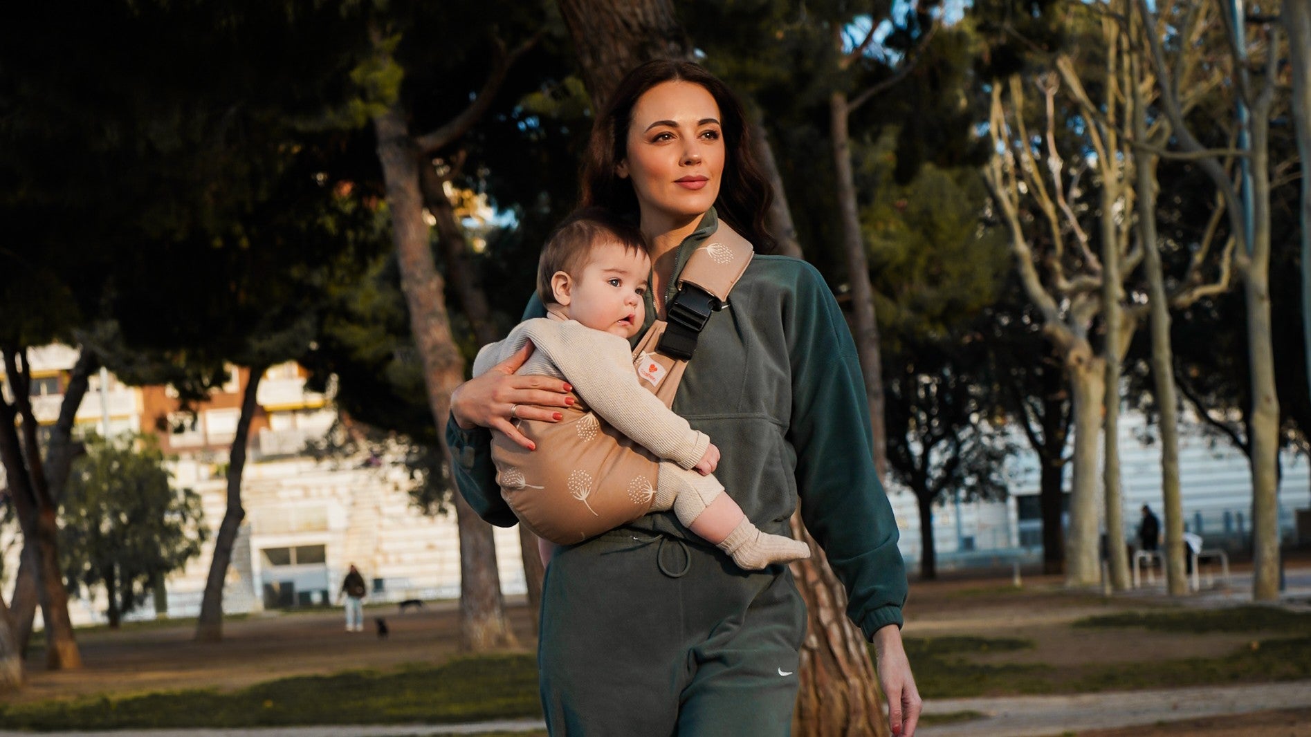 Donna in tuta verde scuro con un marsupio per bambini grandi Coshen color mocha decorato con soffioni, durante una passeggiata nel parco.