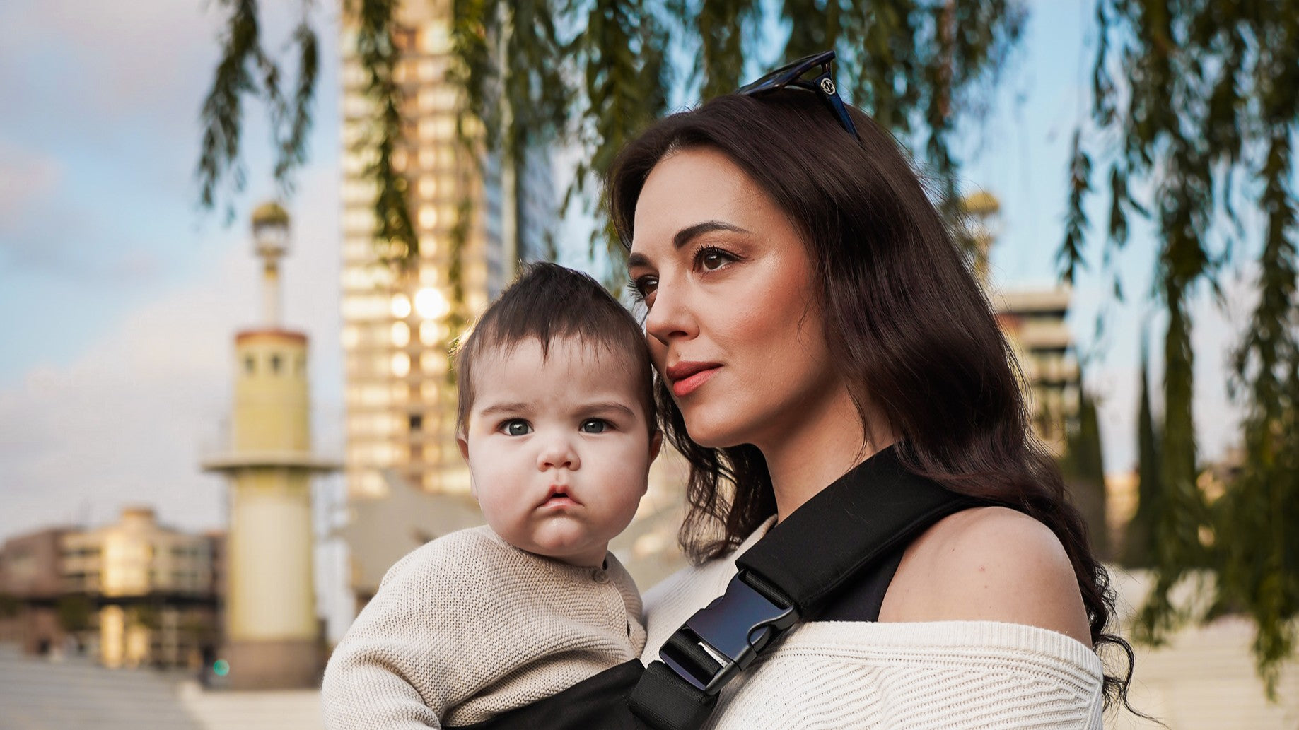 Une femme portant son enfant dans un porte-bébé physiologique noir de la marque Coshen, style urbain chic.