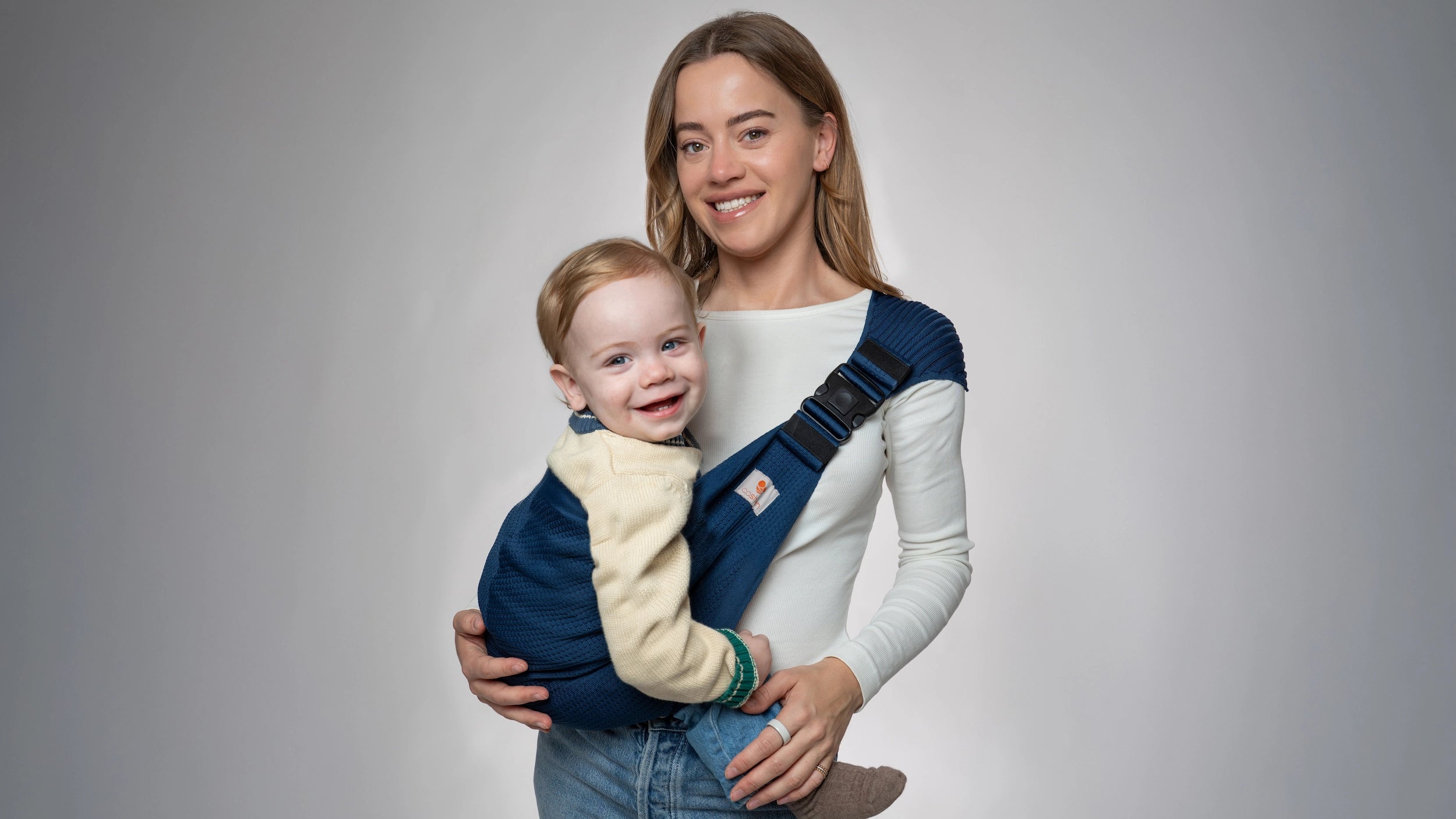 Lachende moeder die haar kind ergonomisch draagt met de Coshen naadloze gebreide peuter heupdrager in donkerblauw; de beste oplossing voor comfortabel en stijlvol babywear op de heup.
