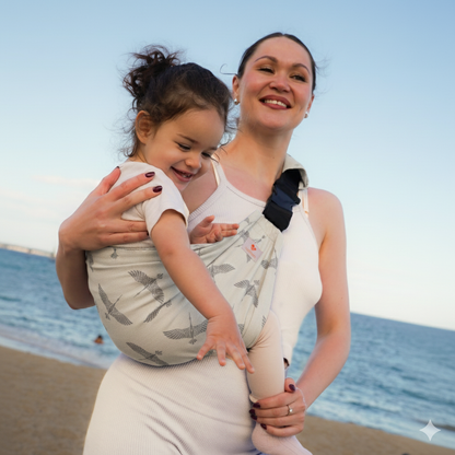 Smiling mother carrying toddler in a leopard print ergonomic baby carrier on the beach at sunset.